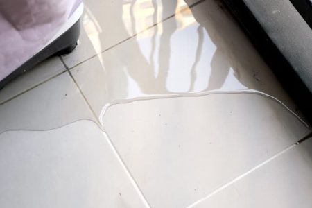 trembling water puddle after pipe burst covers white tile near furniture in room reflecting window and blue sky close view